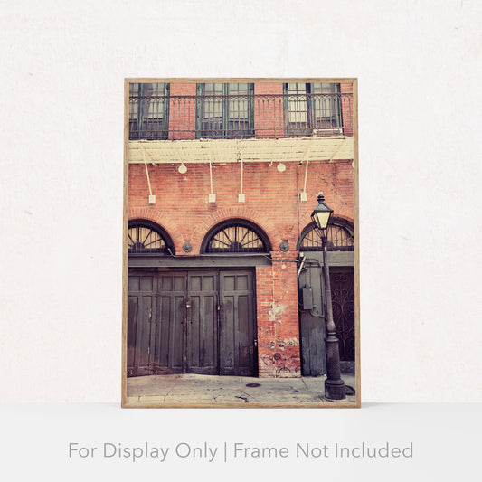 A photography print featuring a vertical composition of an urban scene with a focus on an old, dark door set against a brick wall. There are balcony railings above and a street lamp to the side.