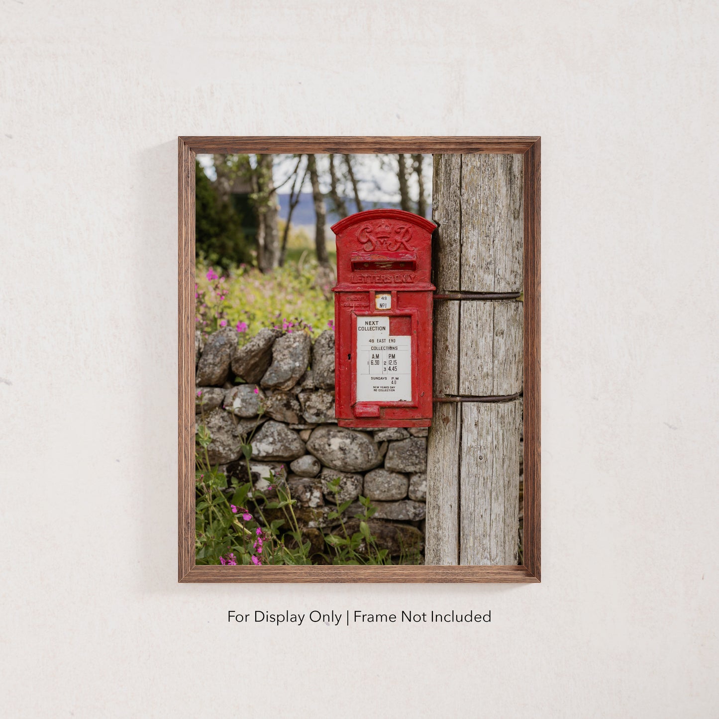 A classic red British post box mounted on a wooden pole beside a stone wall with wildflowers, photographed in soft countryside light.