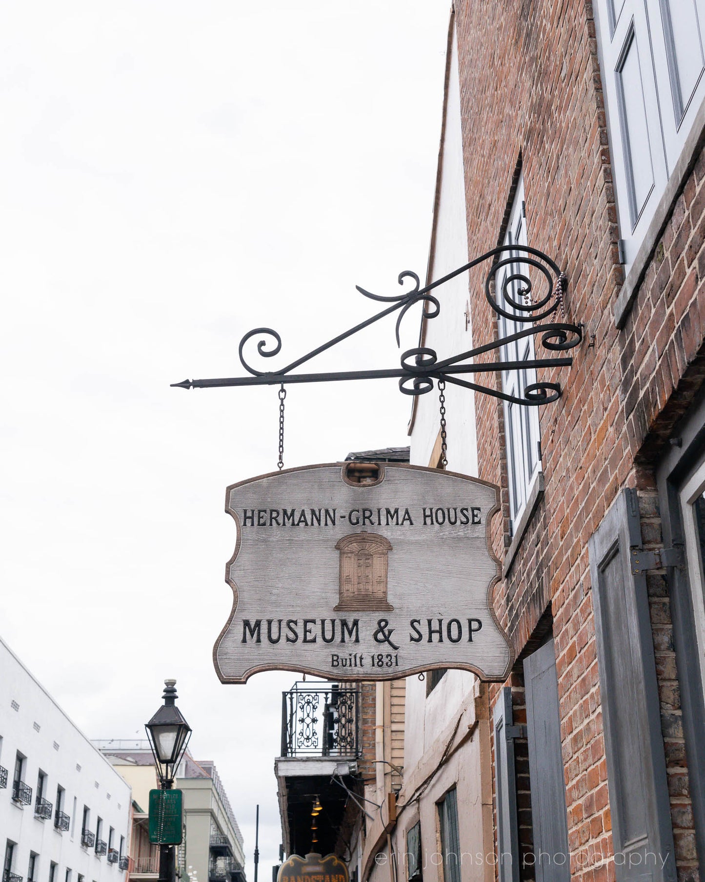 A photograph of a sign for the Hermann-Grima House Museum & Shop, featuring a brick building with an iron scrollwork above the sign.