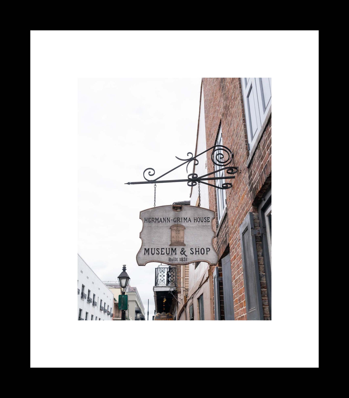 A photograph of a sign for the Hermann-Grima House Museum & Shop, featuring a brick building with an iron scrollwork above the sign.