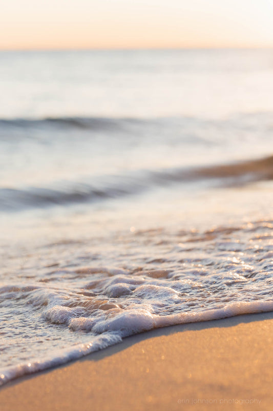 A close-up photograph of a gentle wave lapping against the shore at a beach, with soft sunlight reflecting off the water.