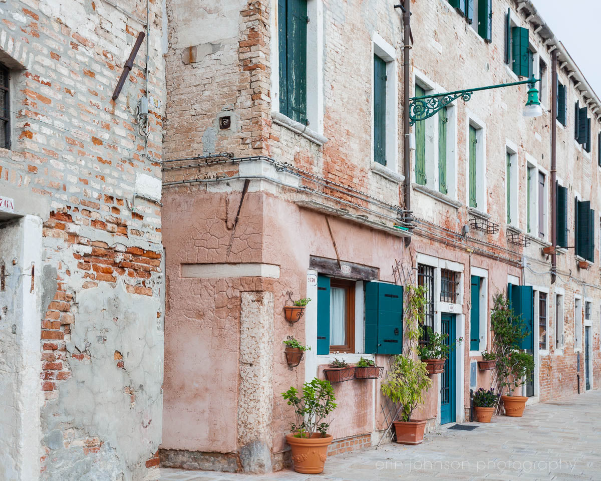 A color photograph of a Venice street showcasing architecture with a focus on old buildings, cobblestone streets, and window boxes with plants.