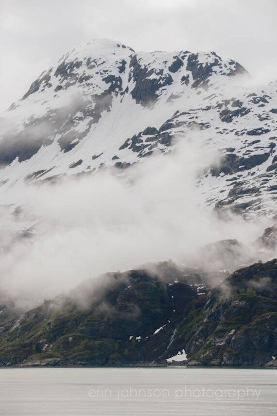 a mountain covered in snow and clouds next to a body of water
