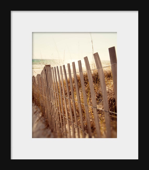A landscape photograph featuring a wooden beach fence, sand dunes, and grass in the foreground, with a serene beach scene.