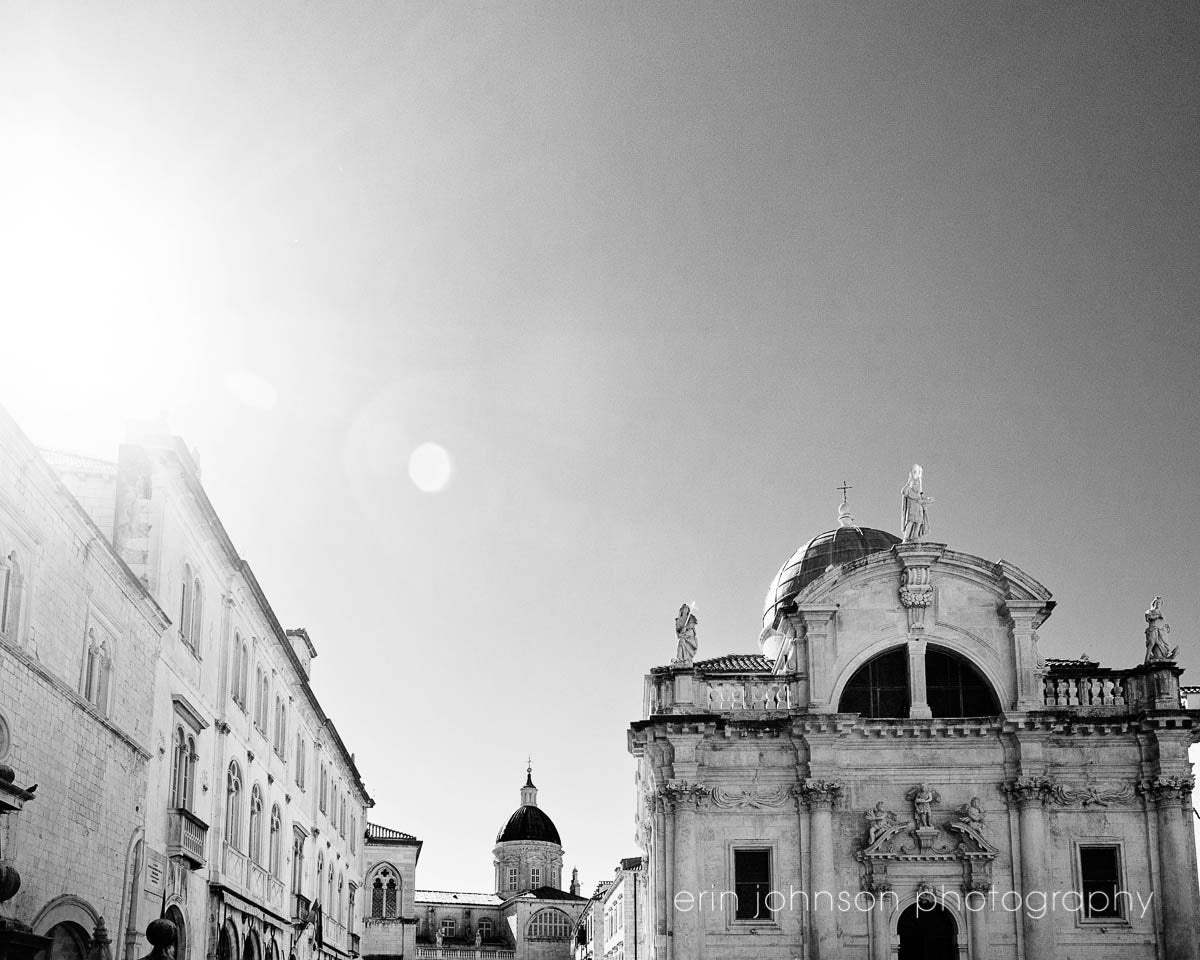 A black and white photograph of a European cityscape in Dubrovnik, Croatia, featuring architecture reminiscent of St. Blaise, with a bright sky background.