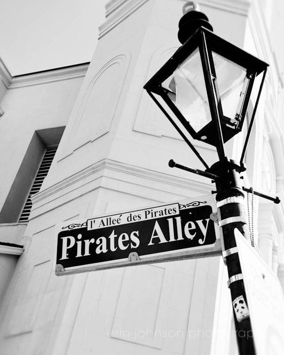 Black and white photograph of a street sign for Pirates Alley with a historical marker, featuring a vintage style lamp post in the background.