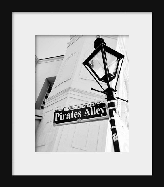 Black and white photograph of a street sign for Pirates Alley with a historical marker, featuring a vintage style lamp post in the background.