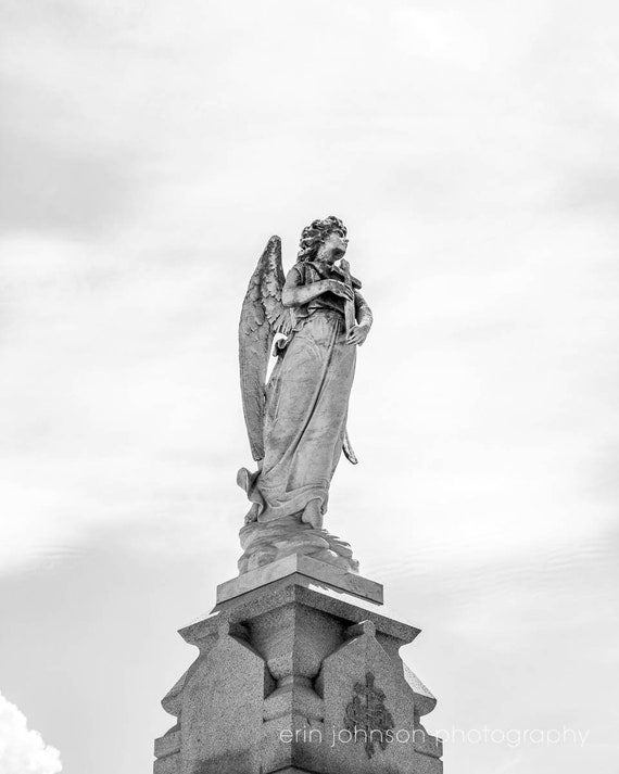 Black and white photograph of an angel statue standing on a pedestal, with wings extended, located in a cemetery setting.