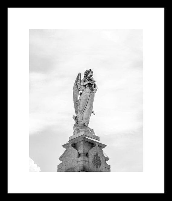Black and white photograph of an angel statue standing on a pedestal, with wings extended, located in a cemetery setting.