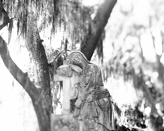Black and white photograph of an angel statue in a garden setting, with a blurred background of trees and foliage.