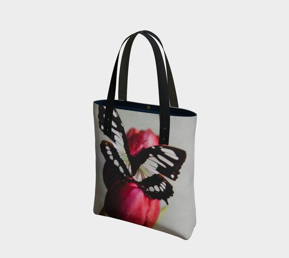 A canvas tote bag with a printed artwork of a butterfly and tulip in a vibrant color palette, featuring black straps and a brass magnetic closure.