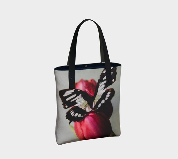 A canvas tote bag with a printed artwork of a butterfly and tulip in a vibrant color palette, featuring black straps and a brass magnetic closure.
