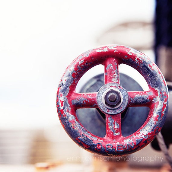 A close-up photograph of a red valve wheel with a blurred industrial background, indicative of an artistic or decorative use.