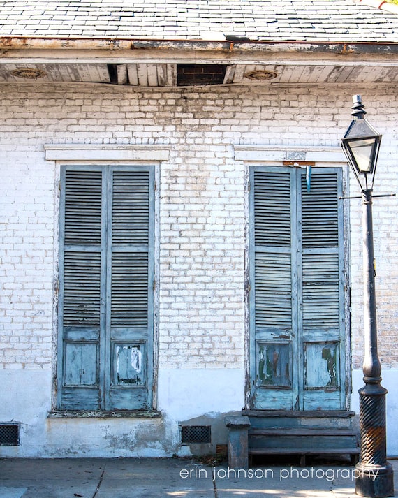 A photograph featuring a pair of green wooden doors with shutters on a white brick wall, illuminated by a lamplight above, taken in a French Quarter setting.