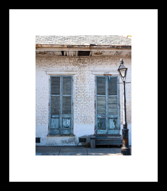 A photograph featuring a pair of green wooden doors with shutters on a white brick wall, illuminated by a lamplight above, taken in a French Quarter setting.