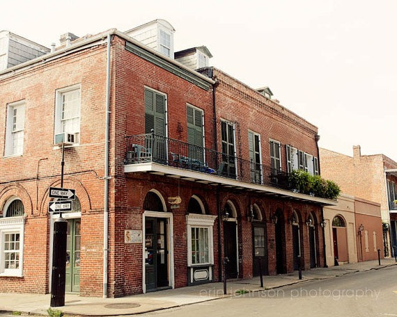 A photograph featuring a brick building with a balcony and a signpost on the side, depicting a street scene in New Orleans.