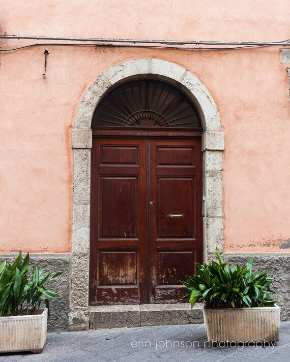 A photograph featuring a pink-tinted doorway on a street in Taormina, Sicily, with a dark stone arch and a wooden door, displayed on a wall.