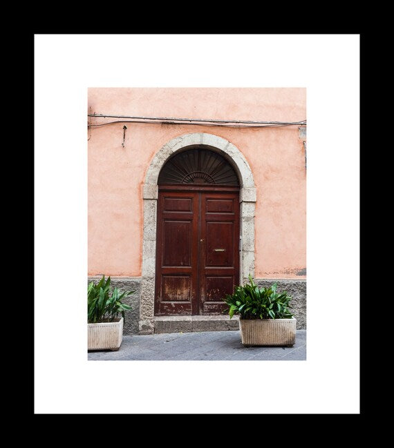 A photograph featuring a pink-tinted doorway on a street in Taormina, Sicily, with a dark stone arch and a wooden door, displayed on a wall.
