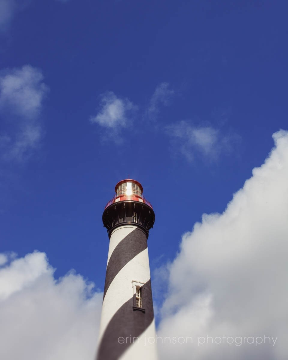 A high-resolution photograph of the Anastasia Island Lighthouse in St. Augustine, Florida, showcasing the lighthouse against a blue sky with clouds.