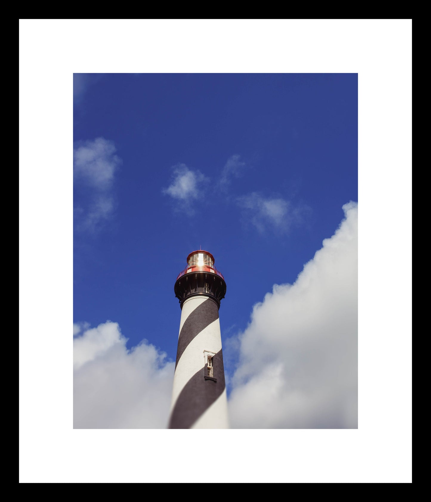 A high-resolution photograph of the Anastasia Island Lighthouse in St. Augustine, Florida, showcasing the lighthouse against a blue sky with clouds.