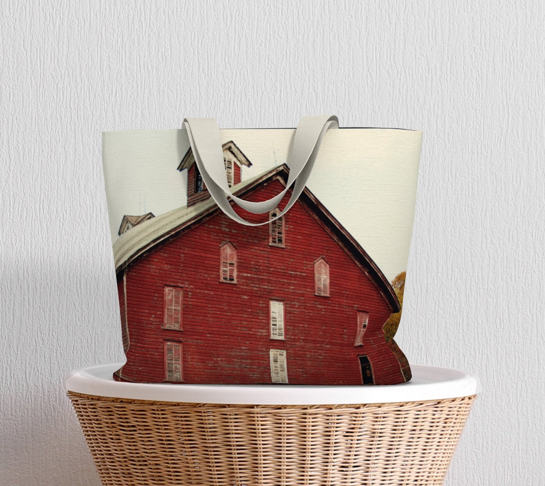A tote bag with a printed rustic barn photography design, featuring a red barn with a navy blue sky background, placed over a wicker basket.