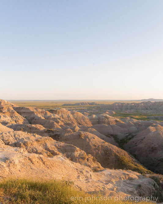A landscape photograph featuring the Conata Basin in South Dakota's Badlands, showcasing a wide-angle view of the flat, arid terrain with distant mountains.
