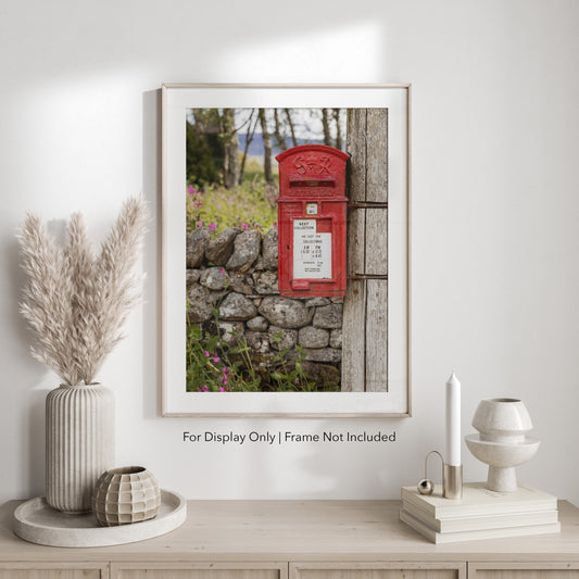 A classic red British post box mounted on a wooden pole beside a stone wall with wildflowers, photographed in soft countryside light.