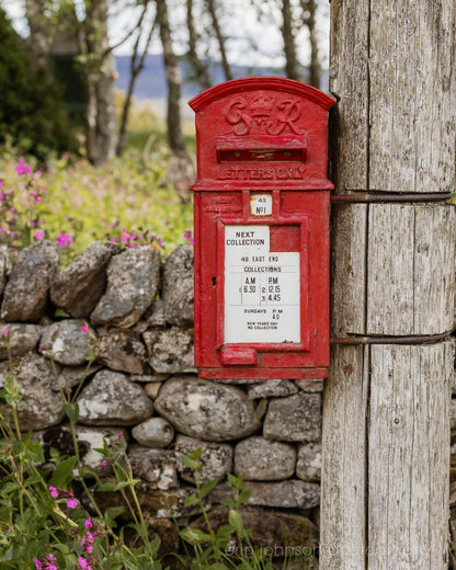 A classic red British post box mounted on a wooden pole beside a stone wall with wildflowers, photographed in soft countryside light.