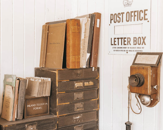 Vintage-style letter box and books on a wooden shelf against a white wall with a 'Post Office' sign.