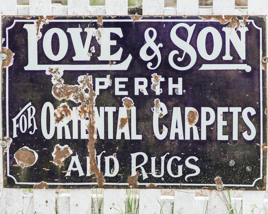 Weathered vintage sign reading “Love & Son Perth for Oriental Carpets and Rugs,” distressed lettering on a rustic black background.