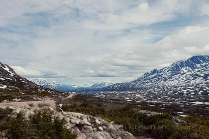 A photography print showcasing a scenic view of the Klondike Highway with mountains in the background, covered in snow.