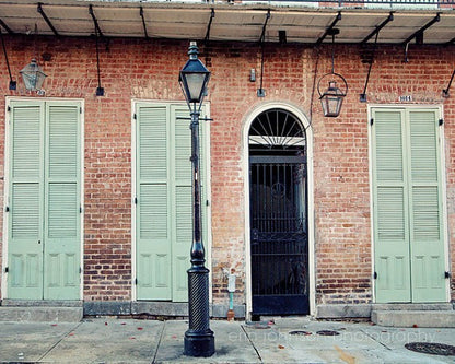 A photograph featuring a black street lamp post with a classic design, in front of a French Quarter building with green doors and windows. New Orleans.